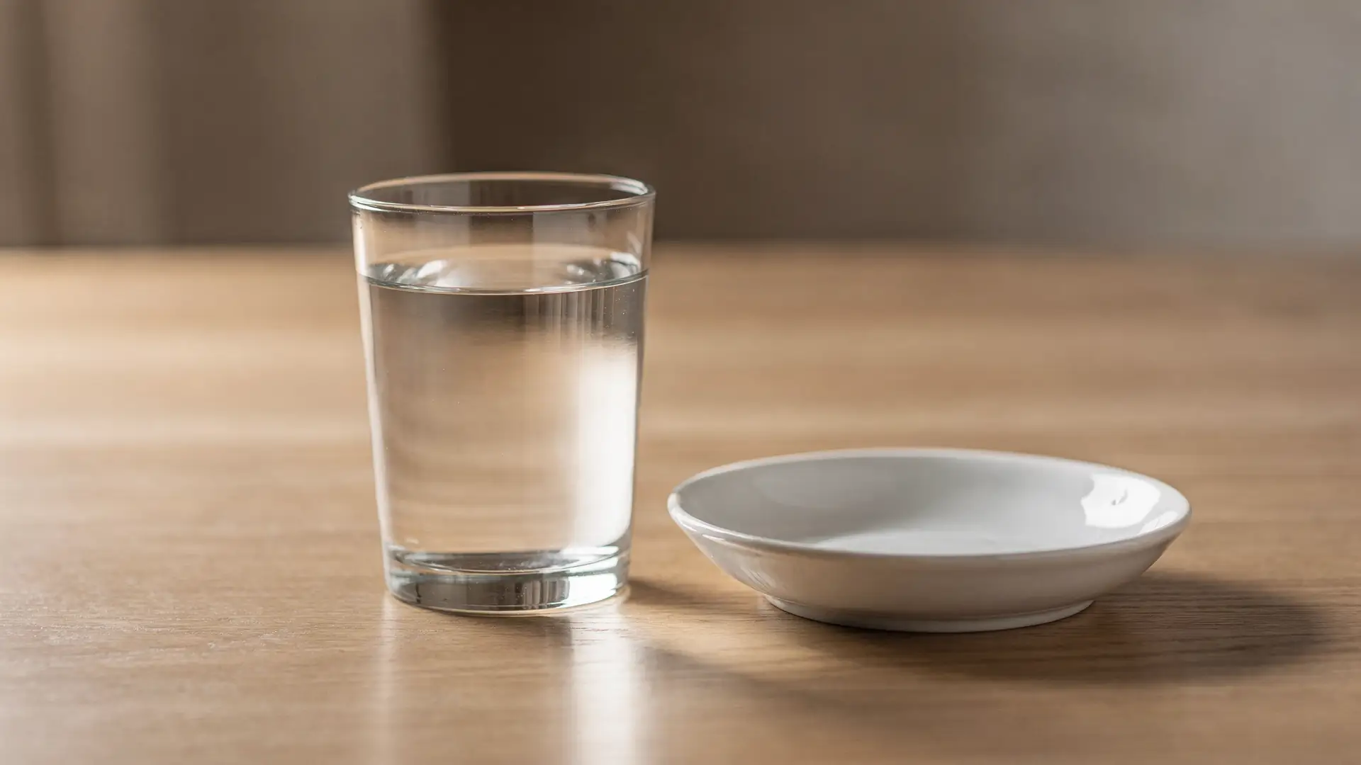 Copo de agua ao lado de pires de ceramica branca vazio sobre mesa de madeira em luz de manha, cena sobre a escolha entre via oral e injetavel