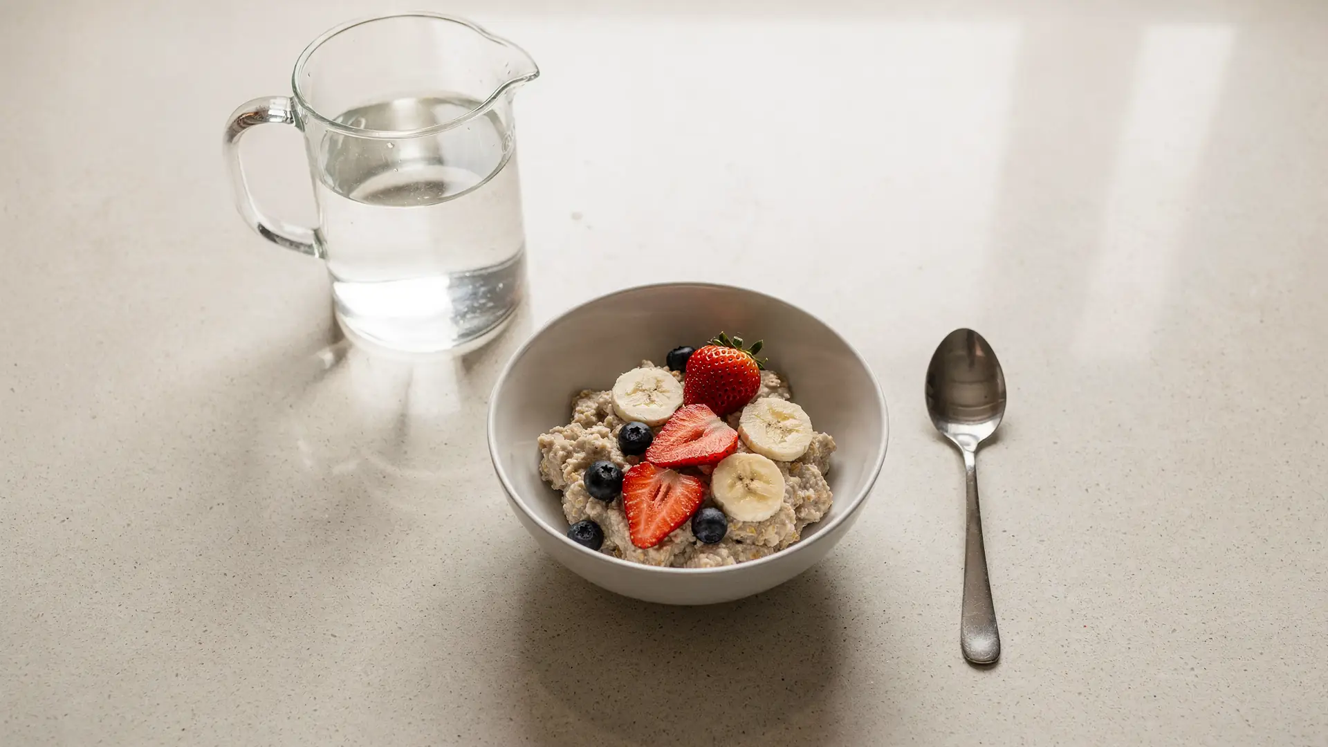 Jarra de agua, prato de aveia com frutas e colher sobre bancada de cozinha clara em luz natural, cena sobre fibra e hidratacao no manejo do intestino durante o tratamento
