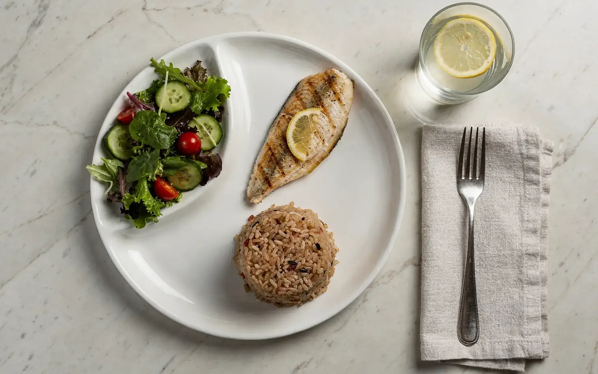 Prato branco visto de cima com salada verde, peixe grelhado e arroz integral, ao lado de um copo de água com limão sobre mesa de mármore claro.