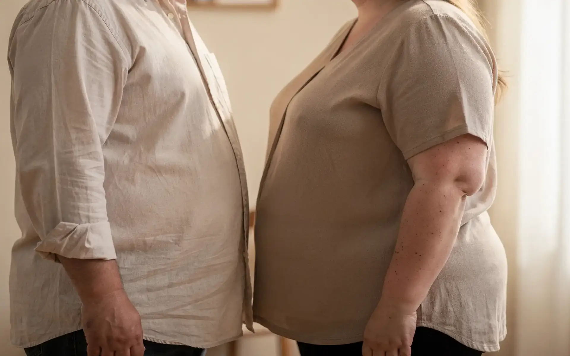 Homem e mulher com obesidade de pé frente a frente, enquadrados do pescoço para baixo, sem rostos no quadro, em ambiente doméstico com luz de manhã, cena que ilustra perda de peso sem motivo.