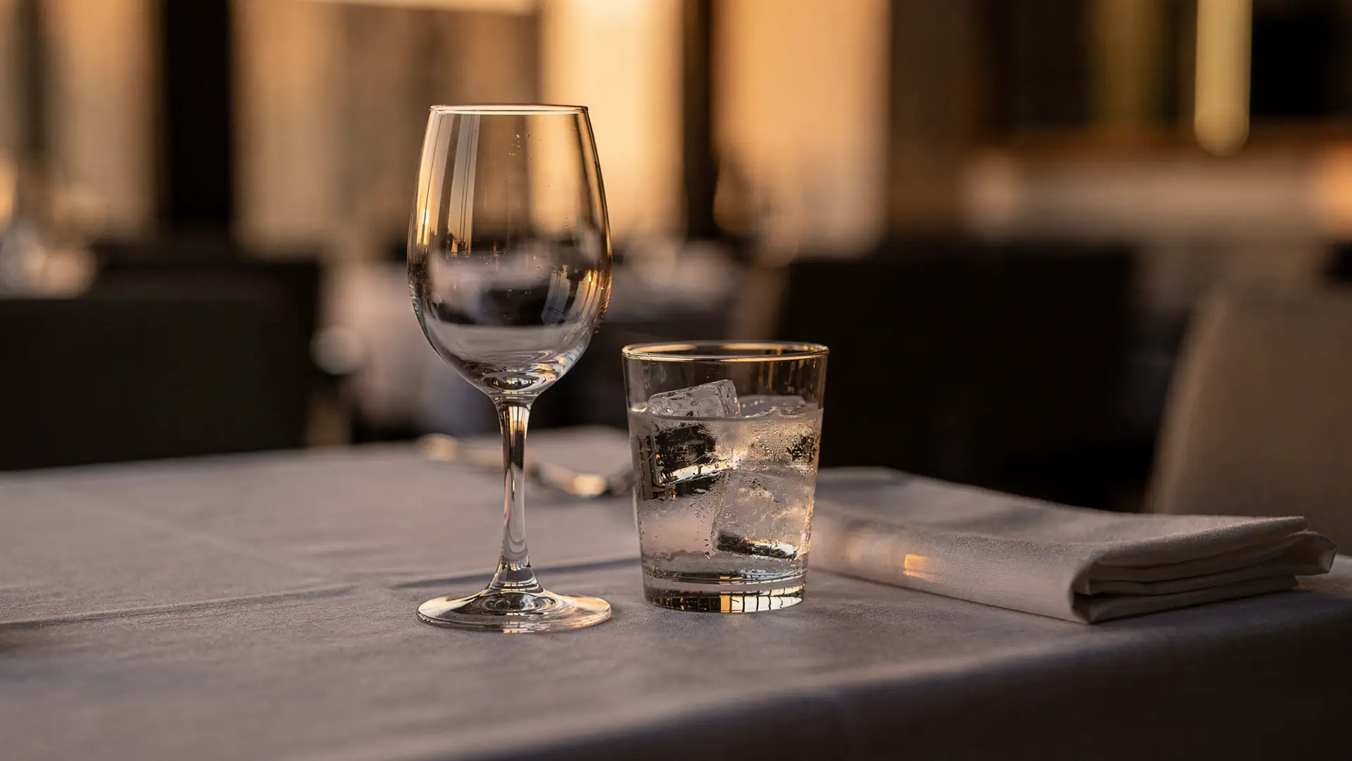 Taca de vinho e copo de agua com gelo sobre mesa posta de restaurante em luz baixa de fim de tarde, cena sobre beber alcool durante o tratamento