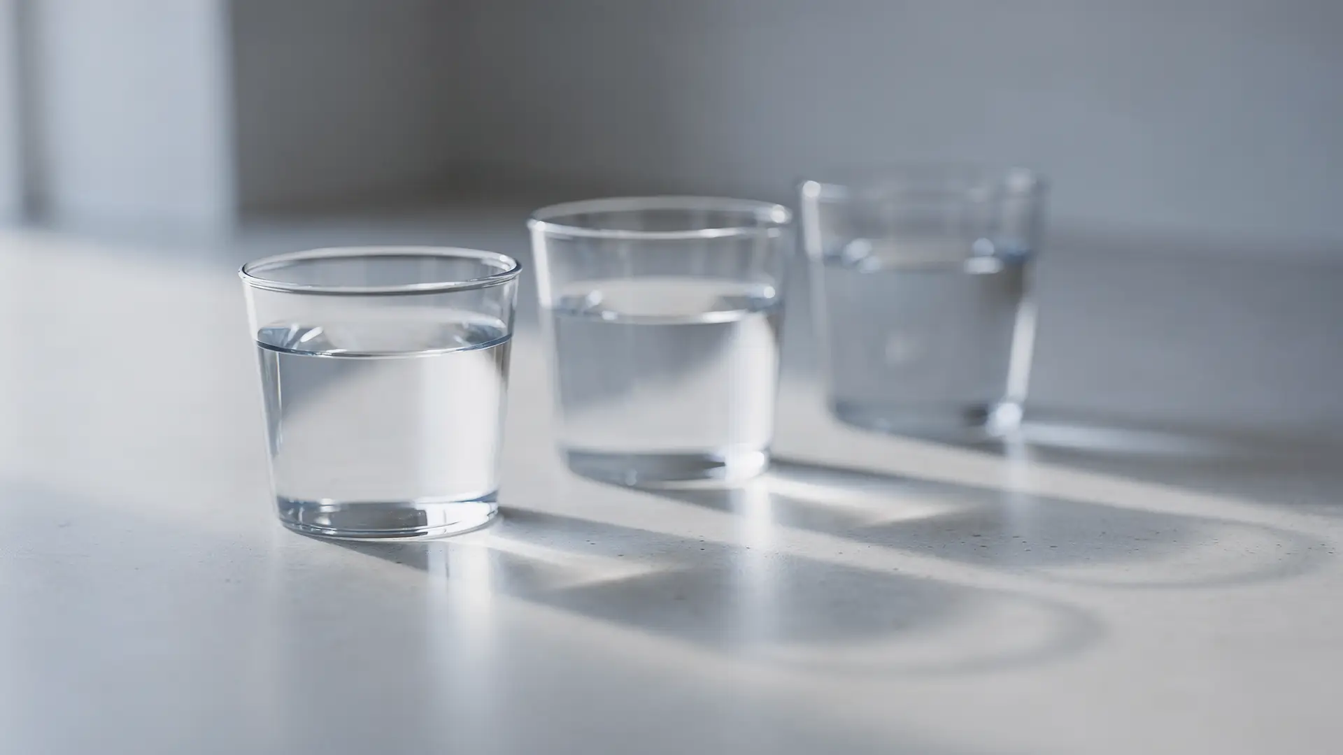 Tres copos de vidro de alturas diferentes com niveis distintos de agua e sombras paralelas sobre bancada clara, cena sobre as diferencas entre as tres substancias